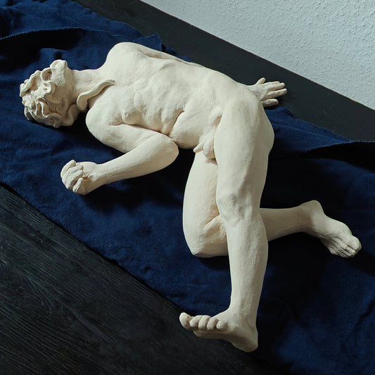 White ceramic sculpture of a reclining figure on a blue cloth against a light gray wall.